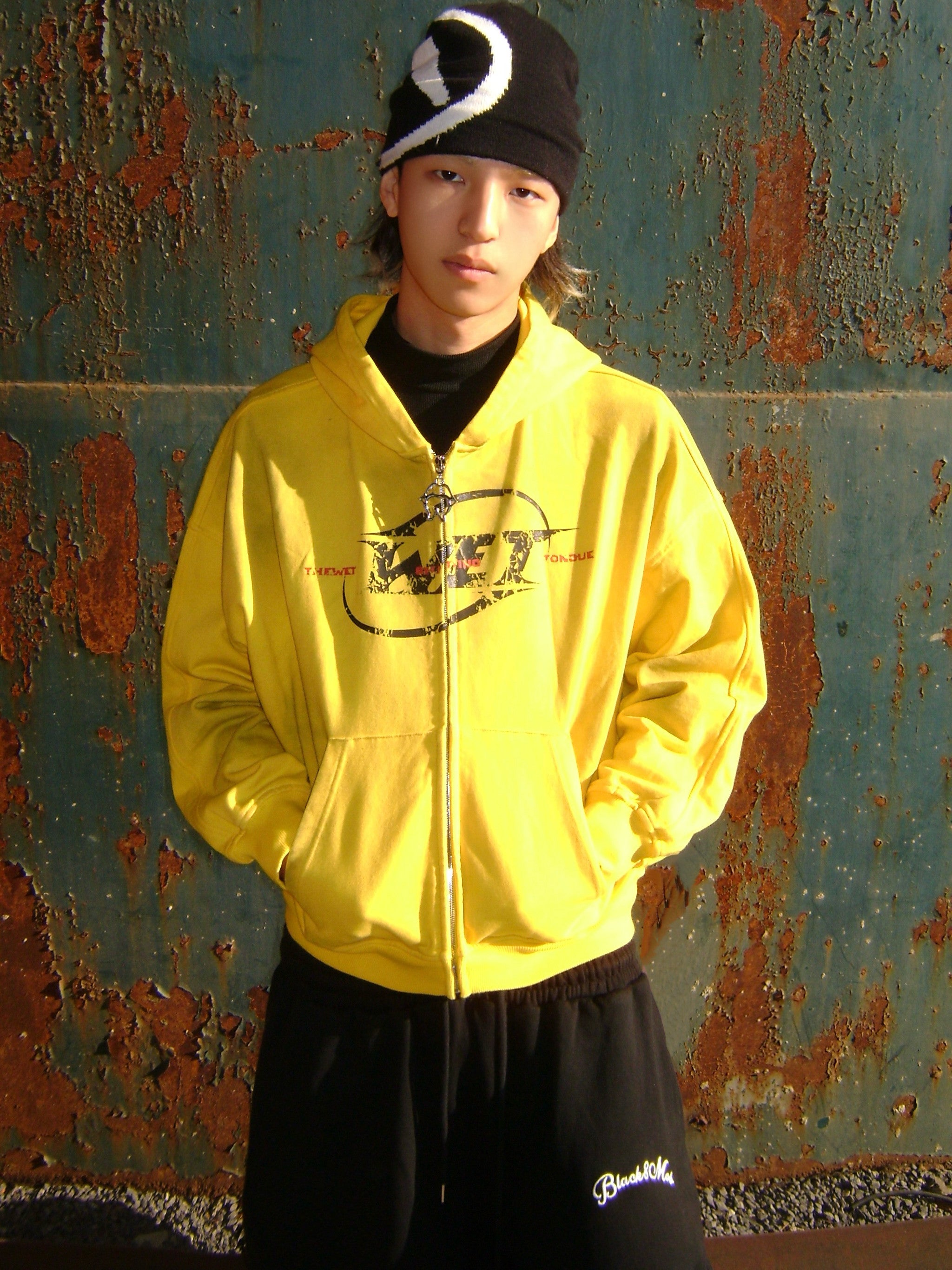 Person wearing a yellow hoodie with a logo in front of a rusty metal wall