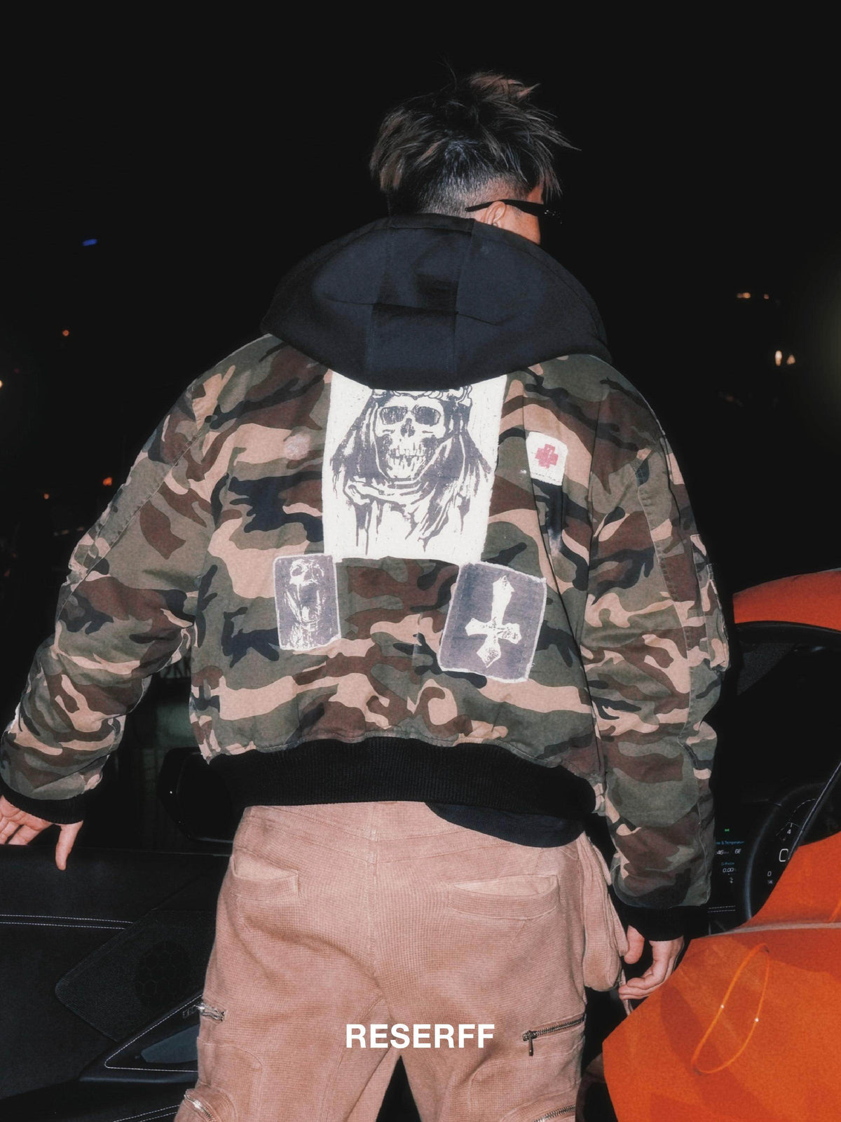 A person viewed from behind wearing a camouflage hoodie with a large skull graphic patch on the back, black hood lining, and two rectangular patch details, paired with tan pants in a dark nighttime setting.