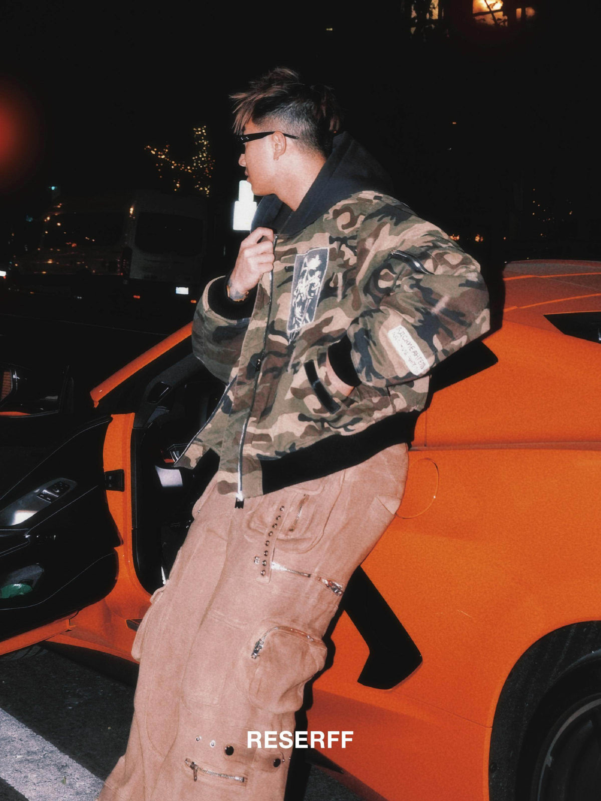 A man wearing a camouflage bomber jacket and beige cargo pants leans against an orange sports car at night. The image has a warm, low-light, grainy aesthetic with a 'RESERFF' watermark at the bottom.